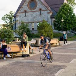 man riding a bike, passing by students sitting on a bench.